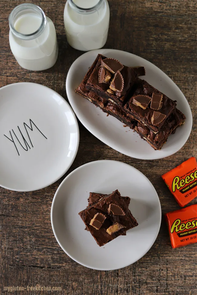 Gluten-free Reeses Brownies on plates with cups of milk and a few Reese's peanut butter cups in package sitting beside