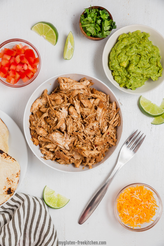 bowl of shredded chicken with bowls of guacamole, tomatoes, and cheese around it