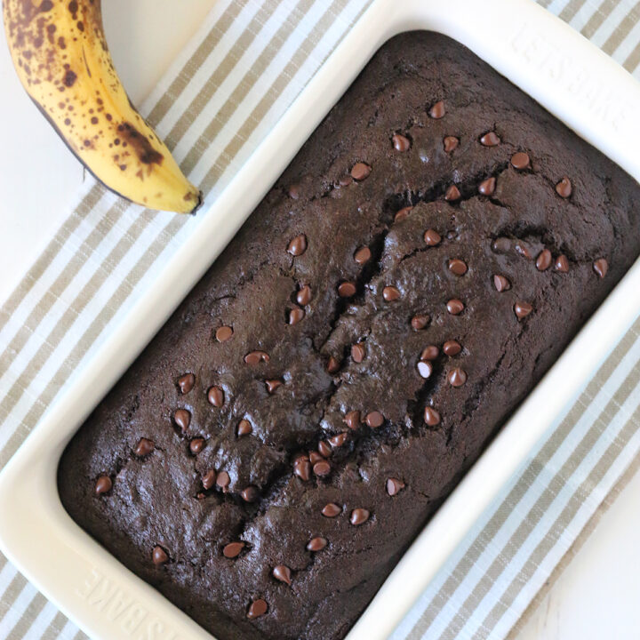 overhead image of loaf of dark brown gluten free chocolate banana bread with miniature chocolate chips in white loaf pan with whole banana next to the pan