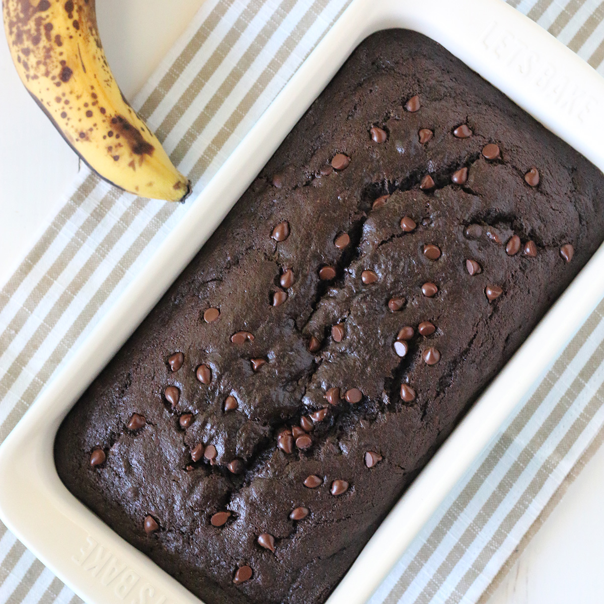overhead image of loaf of dark brown gluten free chocolate banana bread with miniature chocolate chips in white loaf pan with whole banana next to the pan