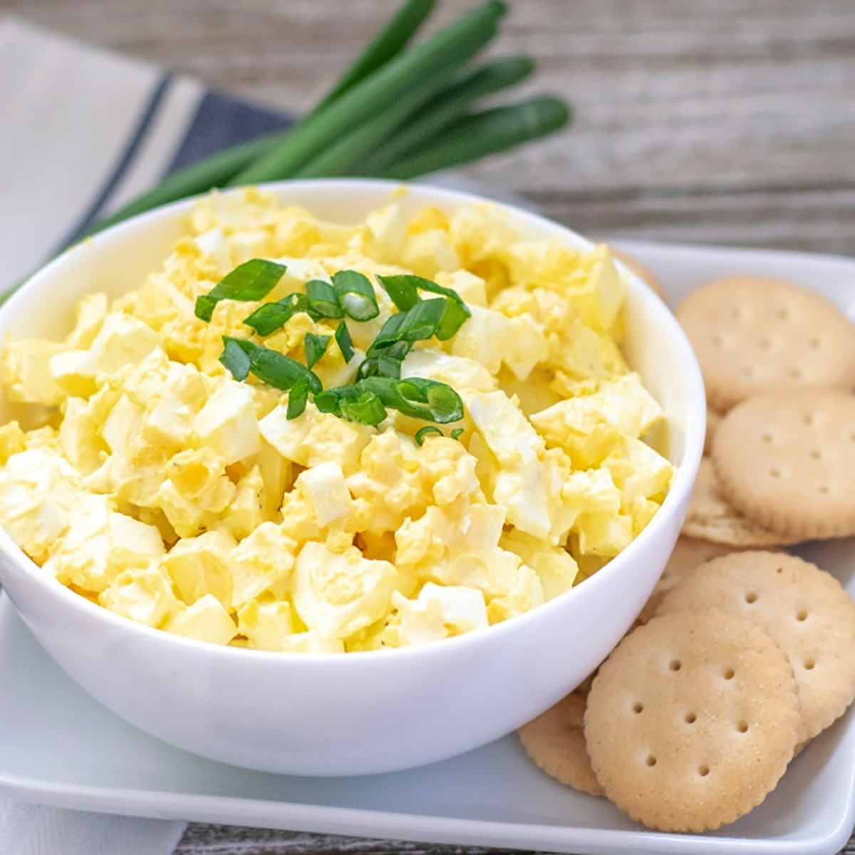 closeup image of white bowl of yellow egg salad with sliced green onions on top and crackers next to it