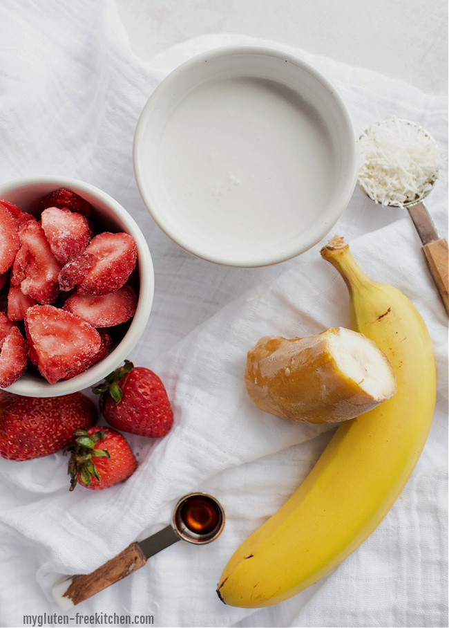 Ingredients for strawberry banana coconut smoothie spread out including bowl of strawberries, one whole banana and a frozen banana, bowl of coconut milk, and some shredded coconut