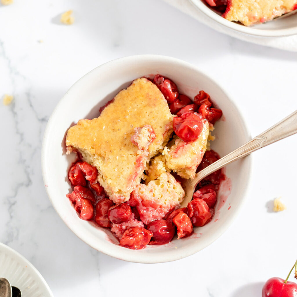 closeup image of Golden cobbler topping and red cherry filling fill a white bowl with a spoon resting inside.