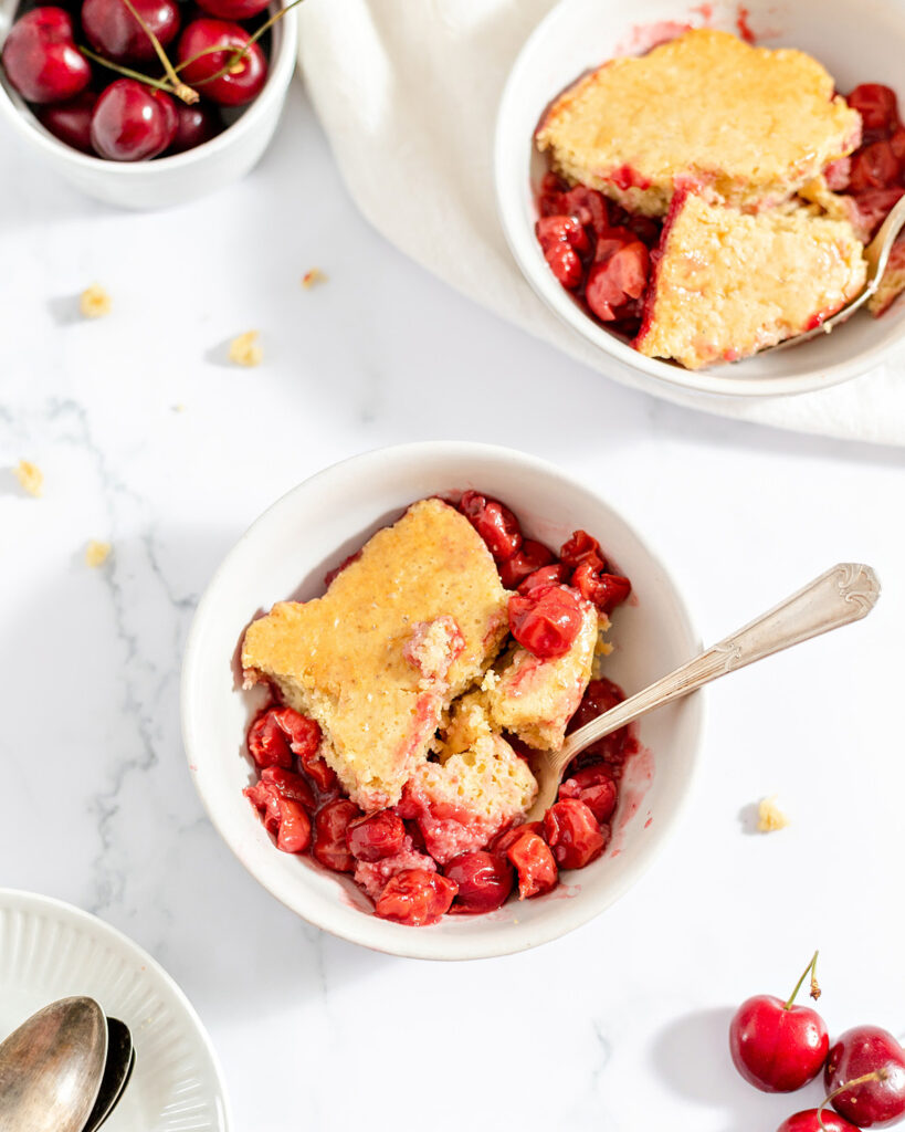 Two bowls of cherry cobbler sit on a light surface with fresh cherries nearby.