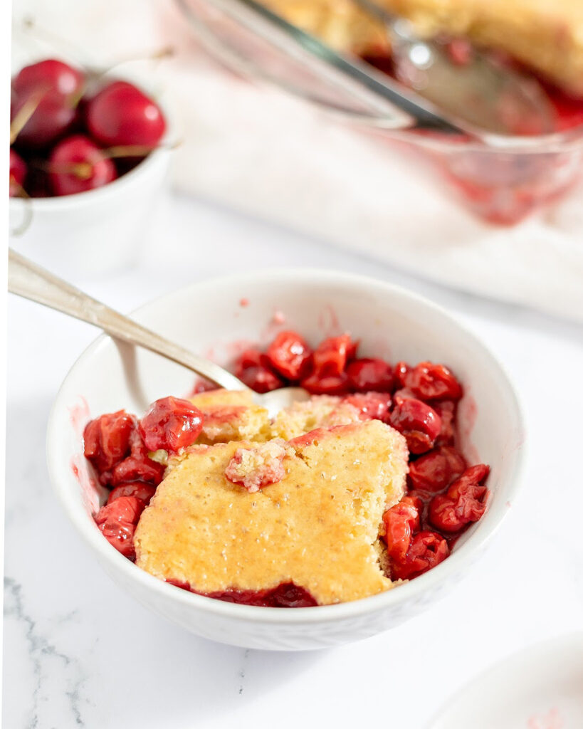 Golden cobbler topping rests over red cherry filling in a white bowl with a spoon.