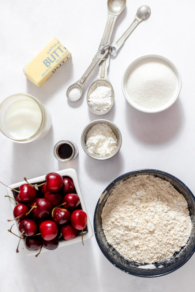 Fresh cherries, gluten free flour, sugar, butter, milk, vanilla, and several white powders are arranged in bowls and measuring spoons on a light surface.