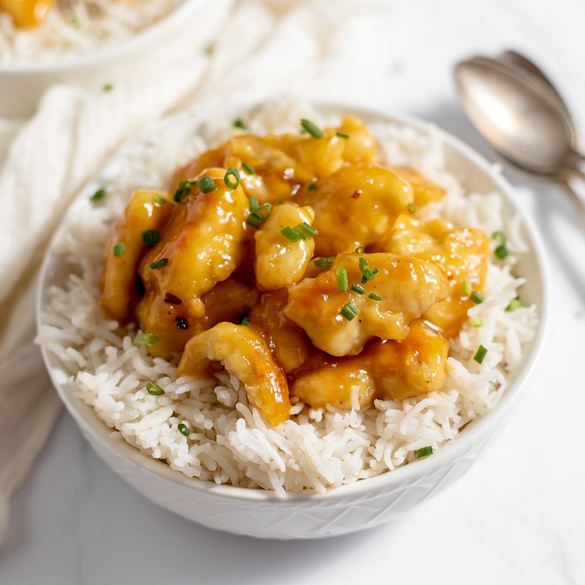 closeup image of orange-glazed chicken pieces resting on white rice with chopped green chives scattered on top.