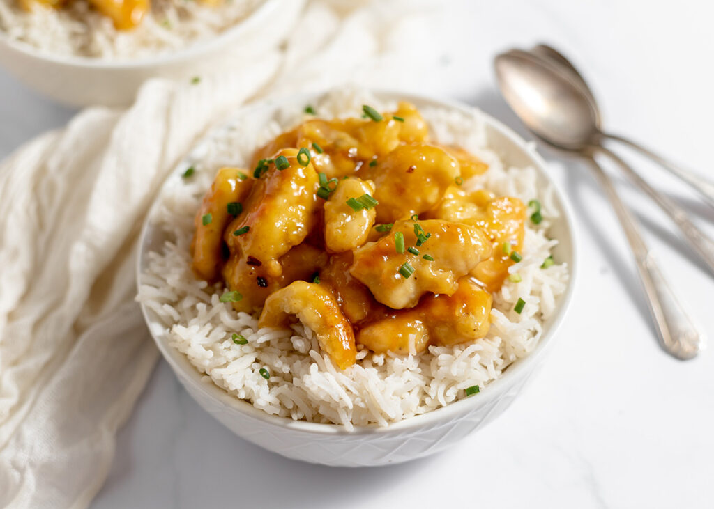A bowl of white rice topped with orange-glazed chicken and chopped green onions sits beside two spoons.