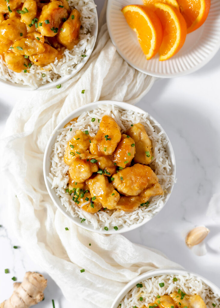 Three bowls of white rice and orange-glazed chicken sit near orange slices, garlic, and ginger.