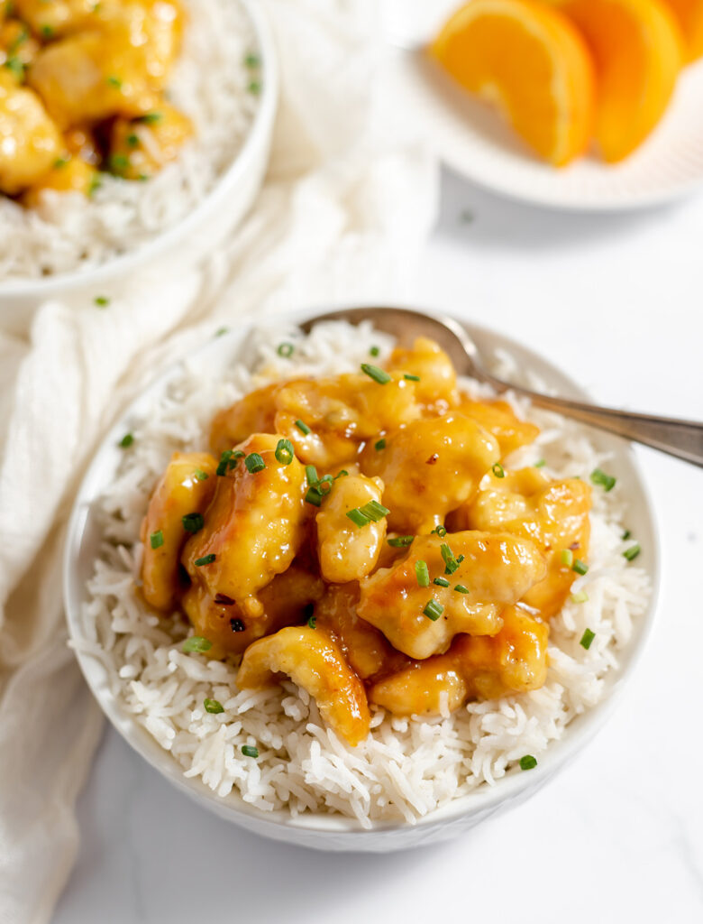 A spoon rests in a bowl of white rice topped with orange-glazed chicken and chopped green chives, with orange slices in the background.