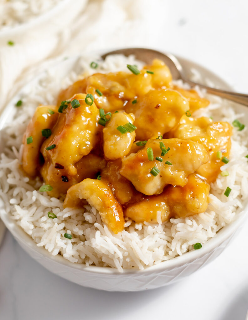 Orange-glazed chicken pieces and chopped green onions top white rice in a white bowl.