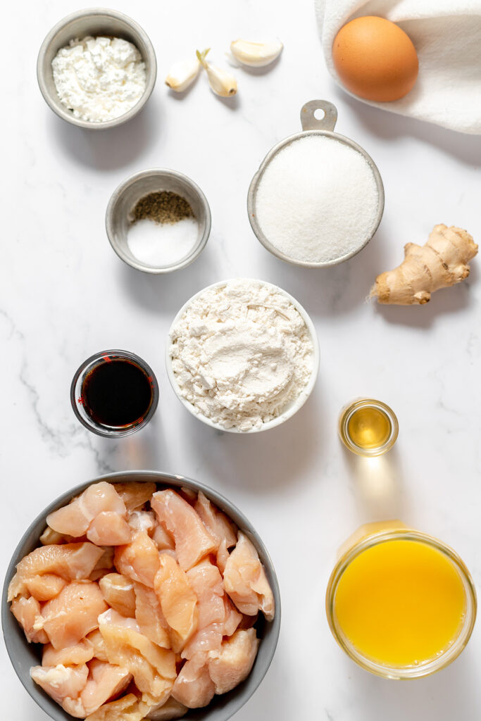 Raw chicken pieces, gluten free flour, cornstarch, sugar, soy sauce, orange juice, olive oil, garlic, ginger, an egg, salt, and pepper are arranged on a light surface.