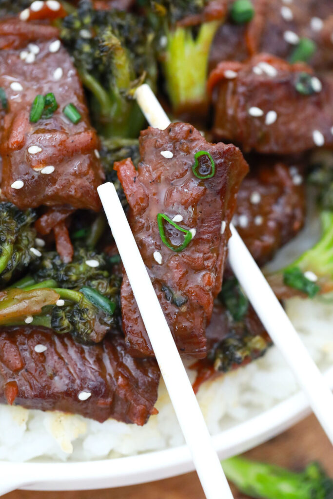 Chopsticks hold a glazed piece of beef over broccoli, rice, and more beef topped with sesame seeds and sliced green onions.