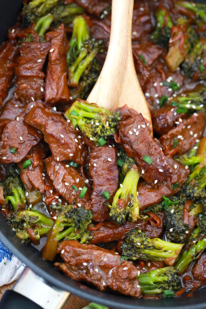 A skillet holds glazed beef and broccoli topped with sesame seeds and sliced green onions, with a wooden spoon in the center.