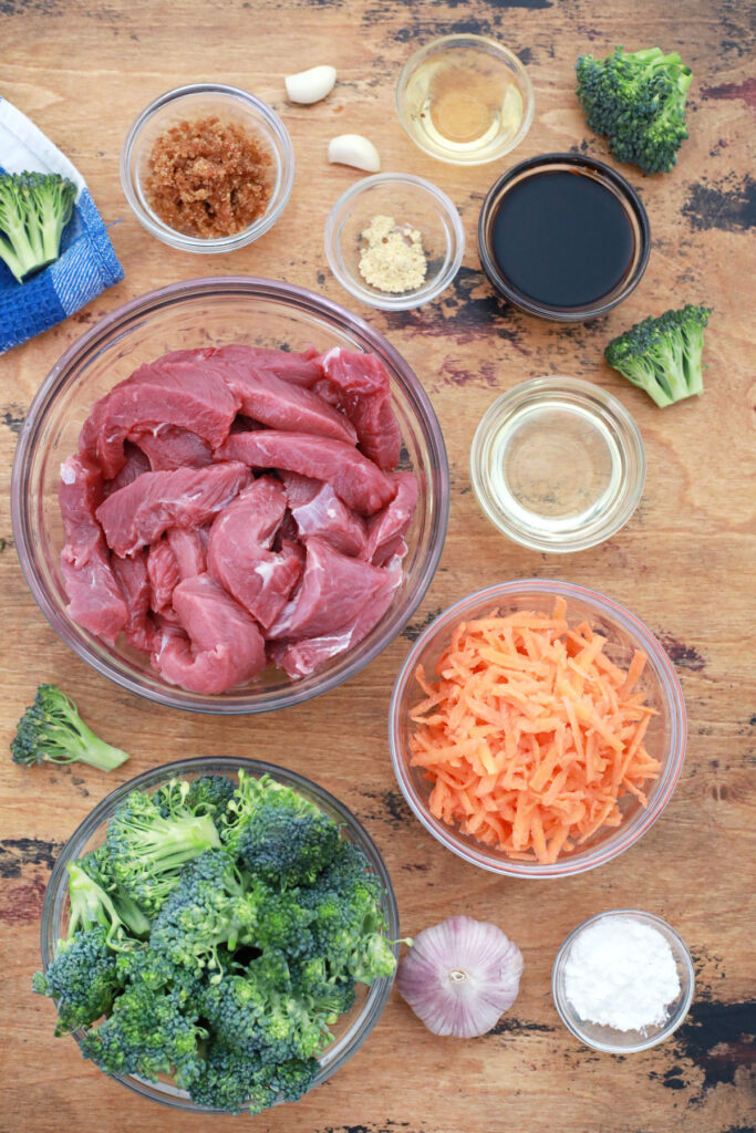 Bowls and dishes hold sliced raw beef, raw broccoli florets, shredded carrots, garlic, dark liquid, clear liquids, brown sugar, and white powders on a wooden surface.