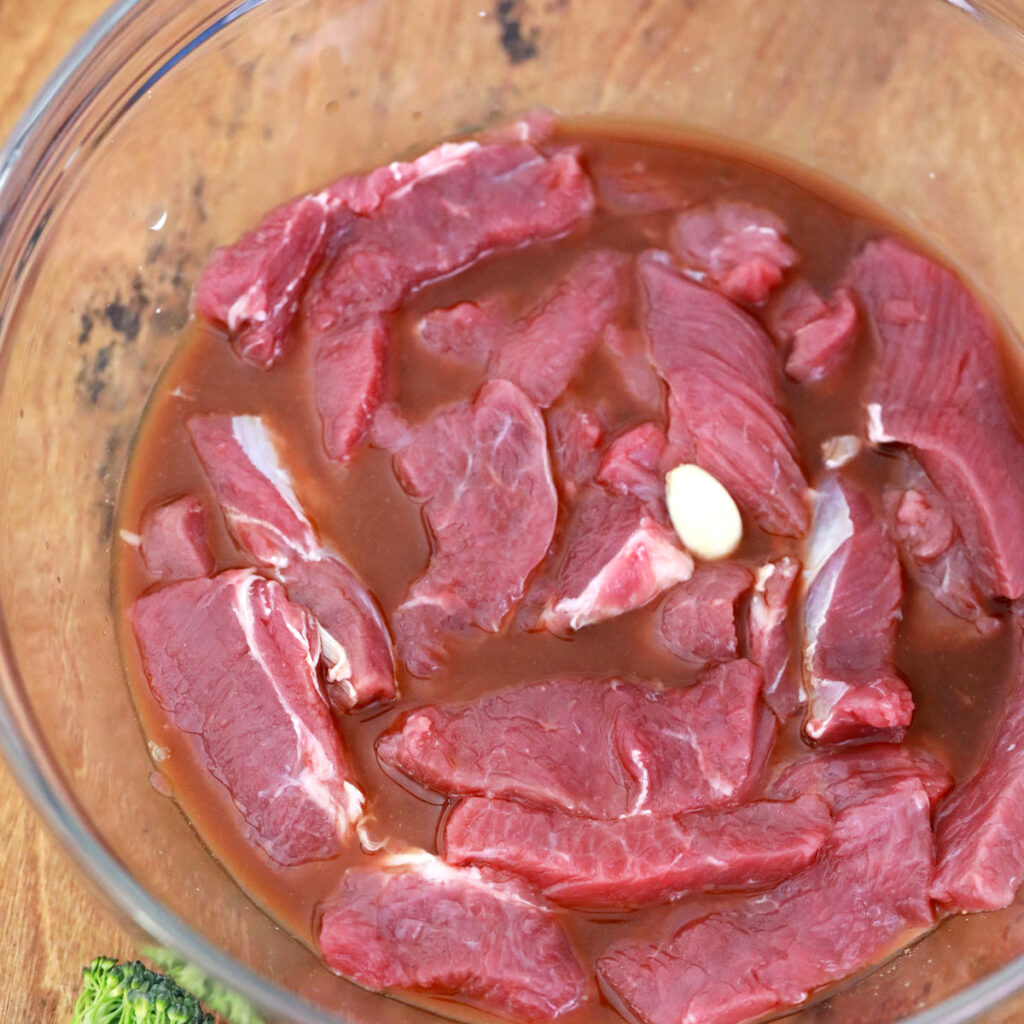 Slices of raw beef soak in a brown liquid with a garlic clove in a glass bowl.