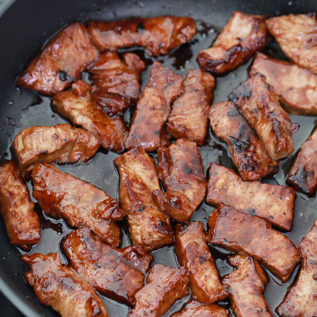 Glazed pieces of beef cook in a dark skillet.