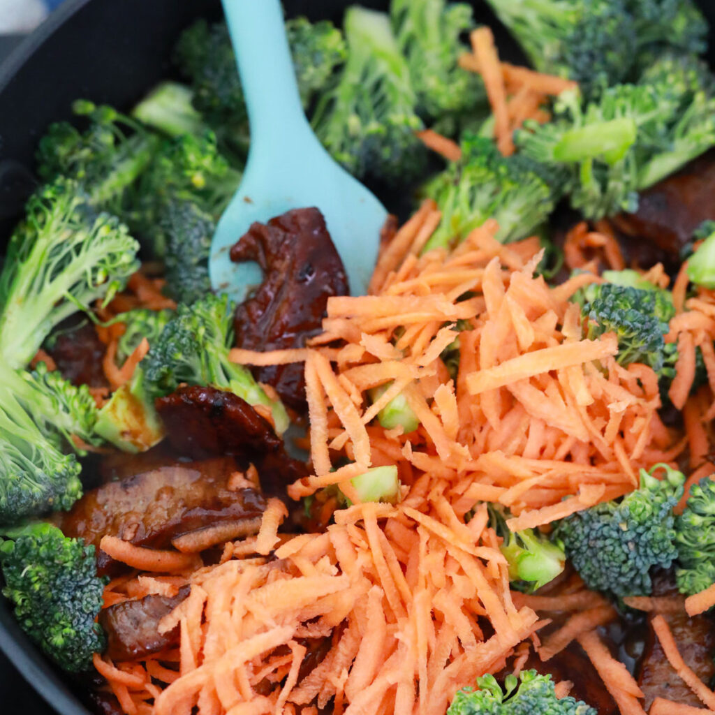 Broccoli florets and shredded carrots sit with glazed beef in a skillet as a blue spatula rests among the ingredients.