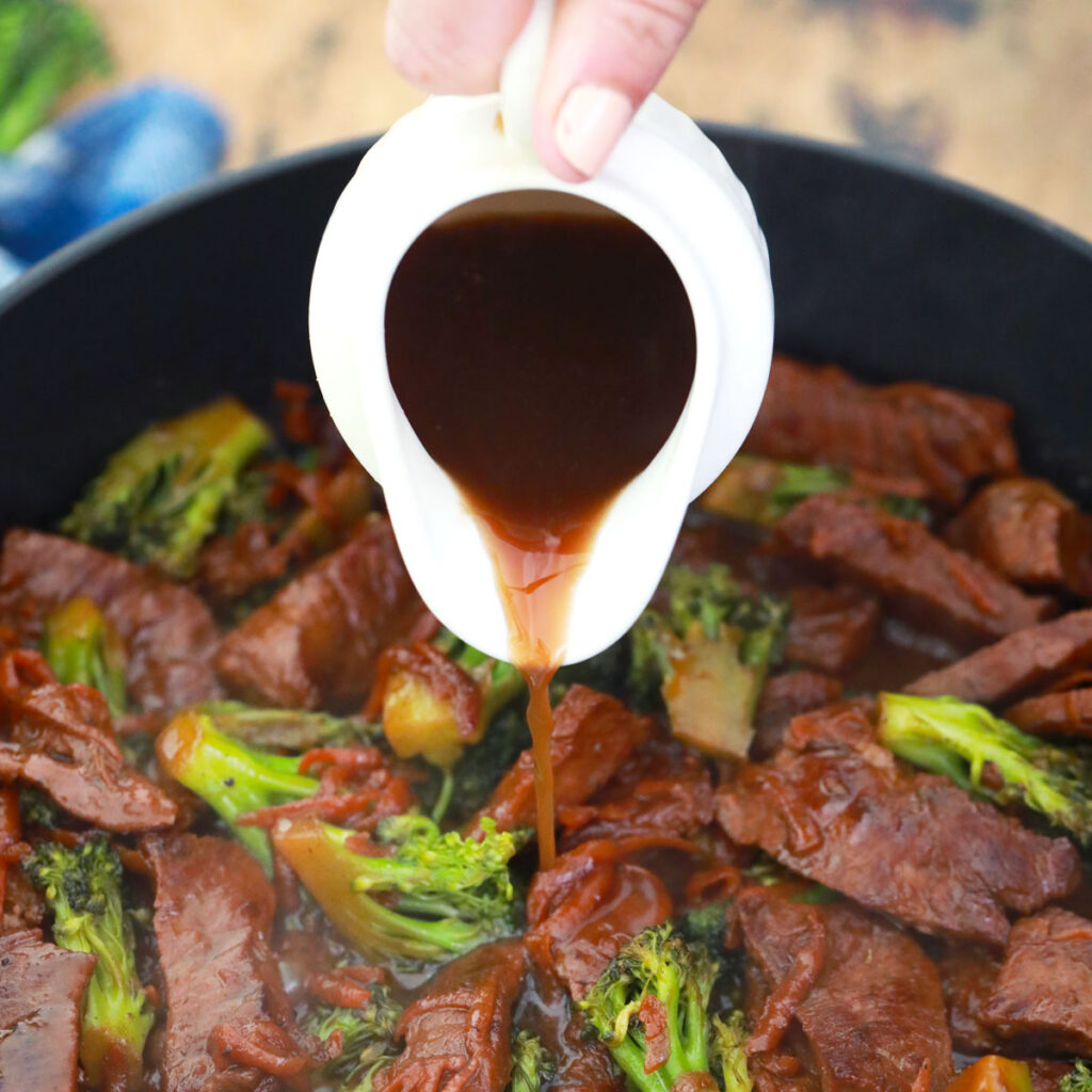 A hand pours brown sauce from a small white pitcher over beef and broccoli in a skillet.