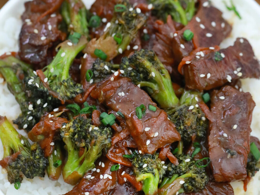 Glazed beef and broccoli topped with sesame seeds and sliced green onions sit over white rice.